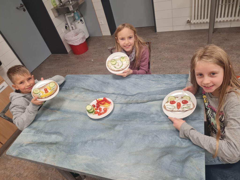 Drei Kinder sitzen an einem Tisch, vor ihnen Teller mit Broten, auf denen mit Gurken, Tomaten und Paprika Gesichter gelegt sind