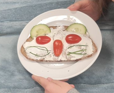 Zwei Hände halten einen Teller, auf dem ein Brot liegt. Das Brot ist mit Tomaten, Gurkenscheiben und Schnittlauch so belegt, dass ich ein katzenartiges Gesicht ergibt