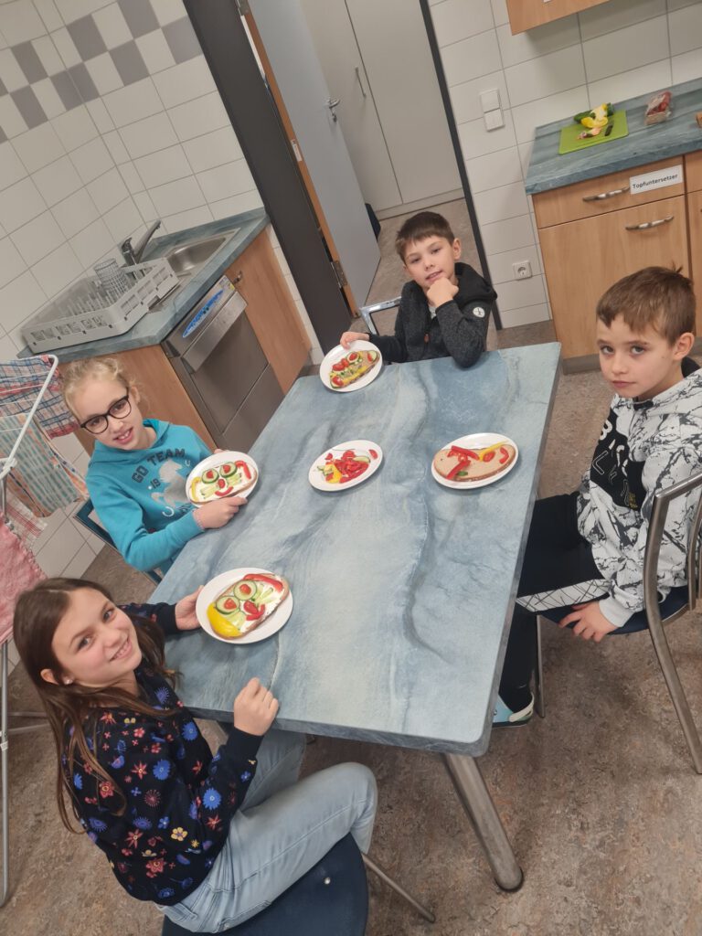 Vier Kinder sitzen an einem Tisch, vor ihnen Teller mit Broten, auf denen mit Gurken, Tomaten und Paprika Gesichter gelegt sind