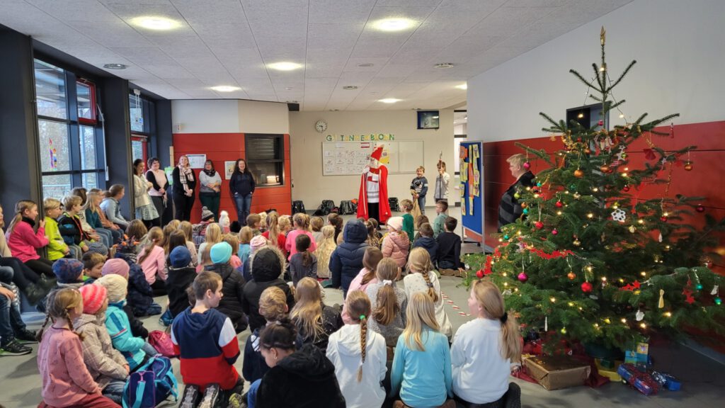 Viele Kinder sitzen in der Pausenhalle auf dem Boden, rechts ist ein geschmückter Weihnachtsbaum zu sehen. Im Hintergrund stehen einige Lehrerinnen neben dem Nikolaus. Ein Kind hält neben dem Nikolaus den Stab, eines hält einen Sack in der Hand.