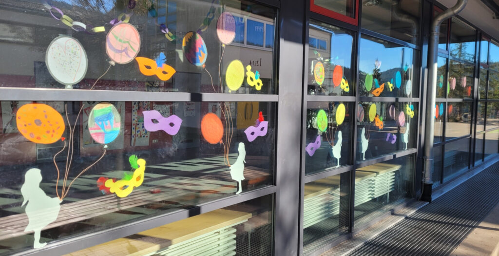 Ein Fensterbild mit mehreren Silhouetten von Mädchen. Mit ihren Händen sind jeweils mit Schnüren Tonpapier-Luftballons verbunden. Dazwischen aus Tonpapier ausgeschnittene Augenmasken.
