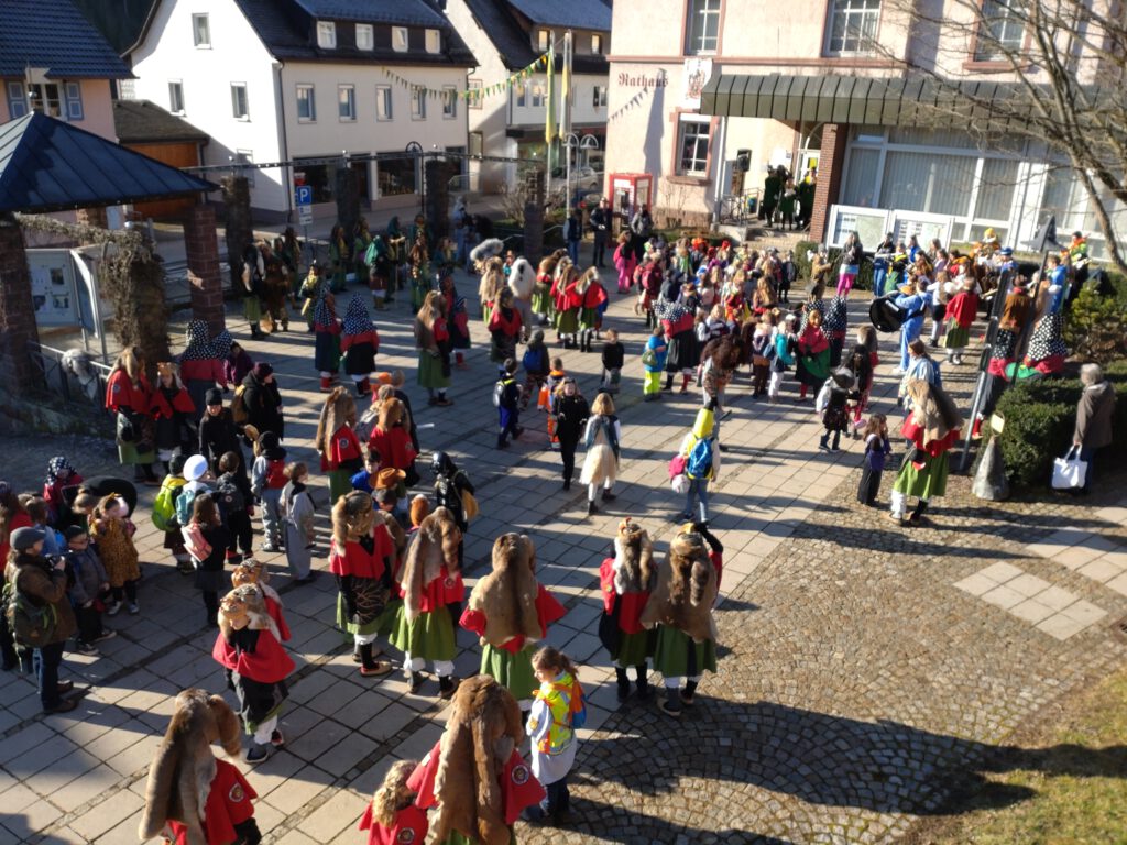 Blick auf den Rathausplatz in Tennenbronn. Vor der Tür des Rathauses stehen einige Menschen in gelb-grünen Kleidern. Auf dem Rathausplatz stehen viele verkleidete Kinder und Vertreter der Tennenbronner Narrenzünfte zusammen