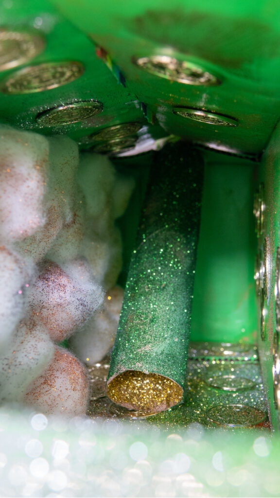 Detailansicht einer gebastelten Falle. Grüner Hintergrund aus Papier, mit Glitzer überzogene Watte, Goldmünzen und eine glitzernd grüne Papprolle