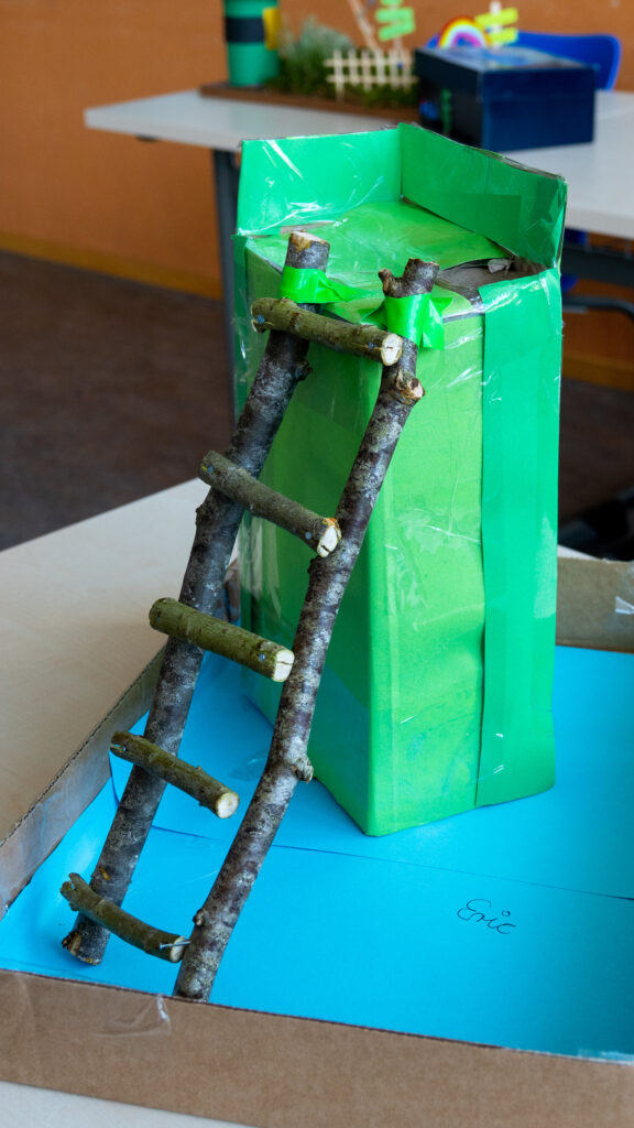 Ein mit grünem Tonpapier beklebter Karton, an dem eine Leiter aus Ästen befestigt ist