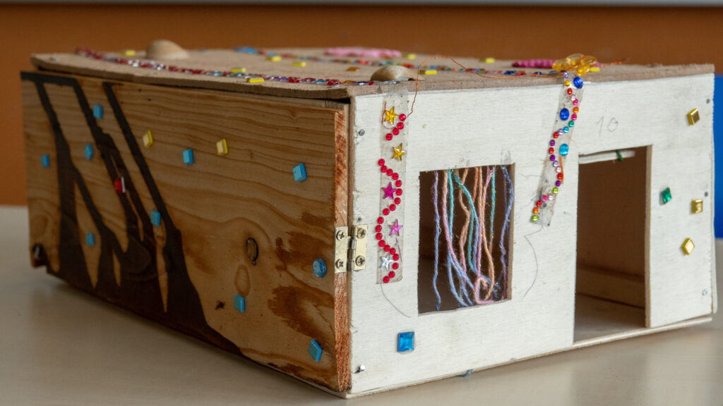 Eine Kiste aus Holz mit einer kleinen, ausgesägten Tür und einem Fenster. Die Kiste ist mit Glitzersteinen und Wolle verziert
