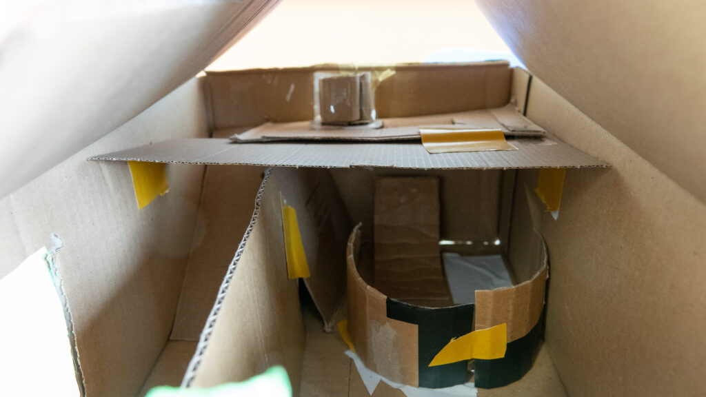 Detailansicht des Inneren einer Pappkiste. Mit Klebeband sind Kartonstreifen zu Gängen, Rampen und Ebenen zusammengefügt