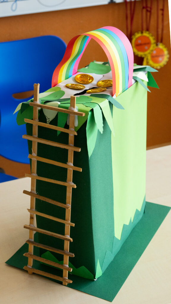 Ein grün beklebter Karton, auf den oben ein Papier-Regenbogen aufgeklebt ist. Auf dem Karton liegen vier Goldmünzen, vorne führt eine aus Holz gebastelte Treppe aufs Kartondach