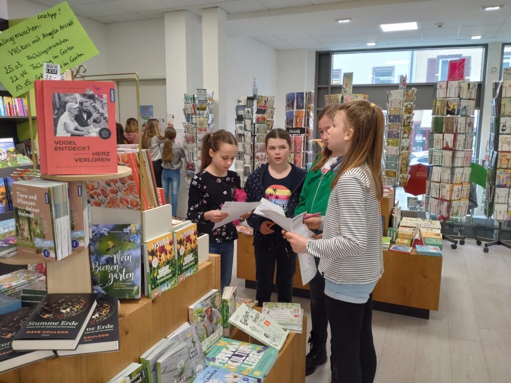 Vier Mädchen stehen um ein niedriges Regal voller Bücher herum und halten Zettel in den Händen