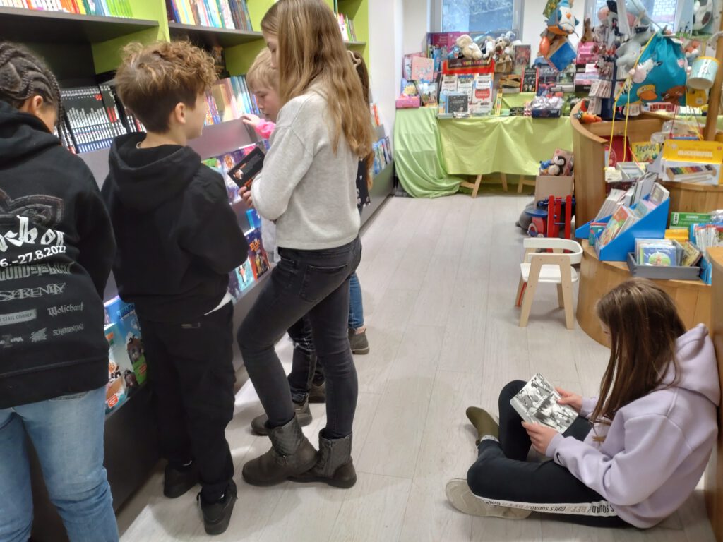 Vier Kinder stehen vor einem Bücherregal und schauen sich die Bücher darin an. Ein Kind sitzt lesend auf dem Boden