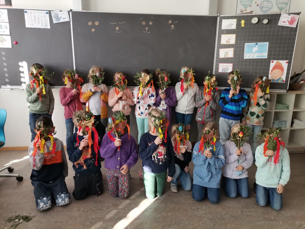 Ein Gruppenbild von Kindern vor einer Tafel im Klassenzimmer. Alle sind zur Kamera gewandt und halten ein mit Palmzweigen und Ostereiern verziertes Kreuz vor ihr Gesicht