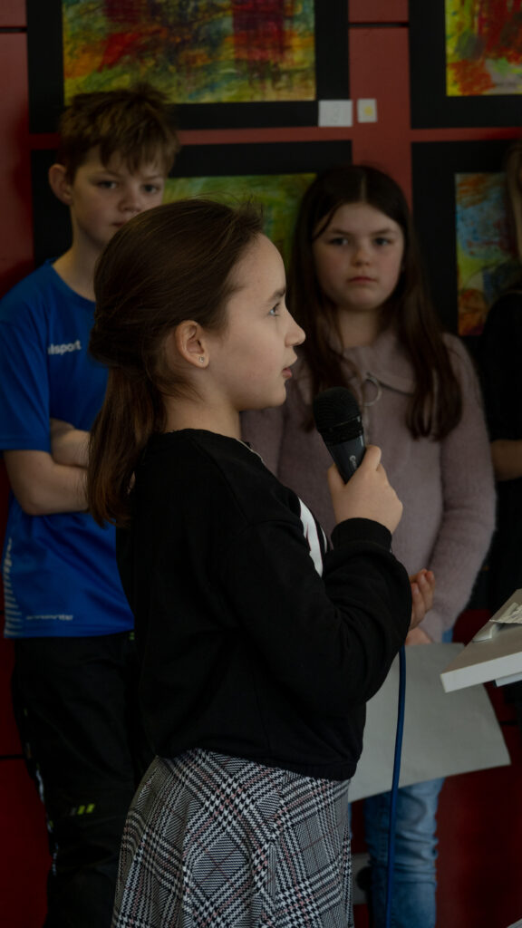 Ein Mädchen, das gerade in ein Mikrofon spricht, ist im Profil zu sehen, im Hintergrund stehen zwei Kinder, die sie anblicken