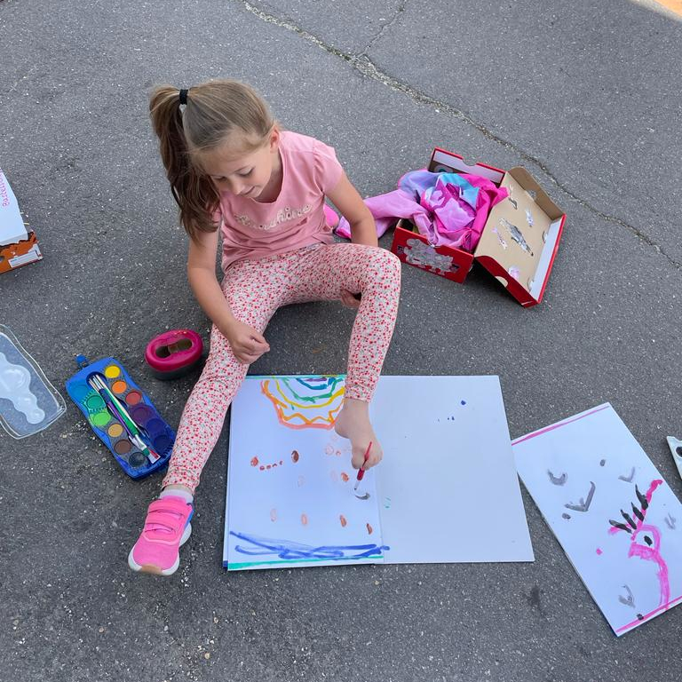 Ein Mädchen sitzt im Schulhof auf dem Boden, vor ihr liegt ein Zeichenblock, neben ihr ein Wasserfarbkasten. Sie hat einen Pinsel zwischen die Zehen geklemmt und malt damit auf dem Zeichenblock einen Regenbogen