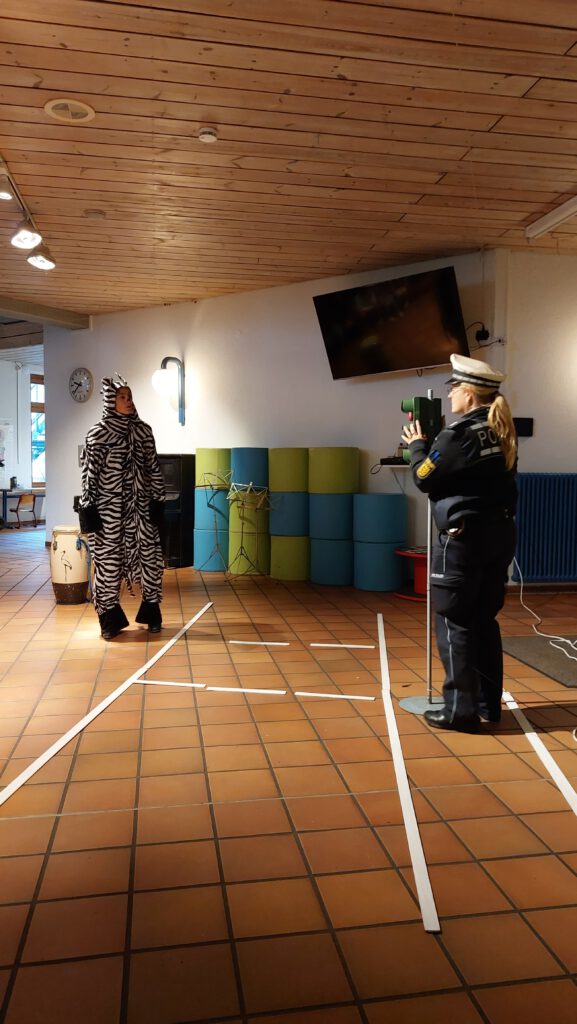 Eine Polizistin steht hinter einer kameraähnlichen Apparatur, die auf eine als Zebra verkleidete Frau gerichtet ist. Auf dem Boden ist mit Papierstreifen eine Straße angedeutet