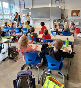 Die Kinder sind im Klassenzimmer an ihren Tischen sitzend von hinten zu sehen. An der Tafel steht Janeth Madrid und erzählt etwas.