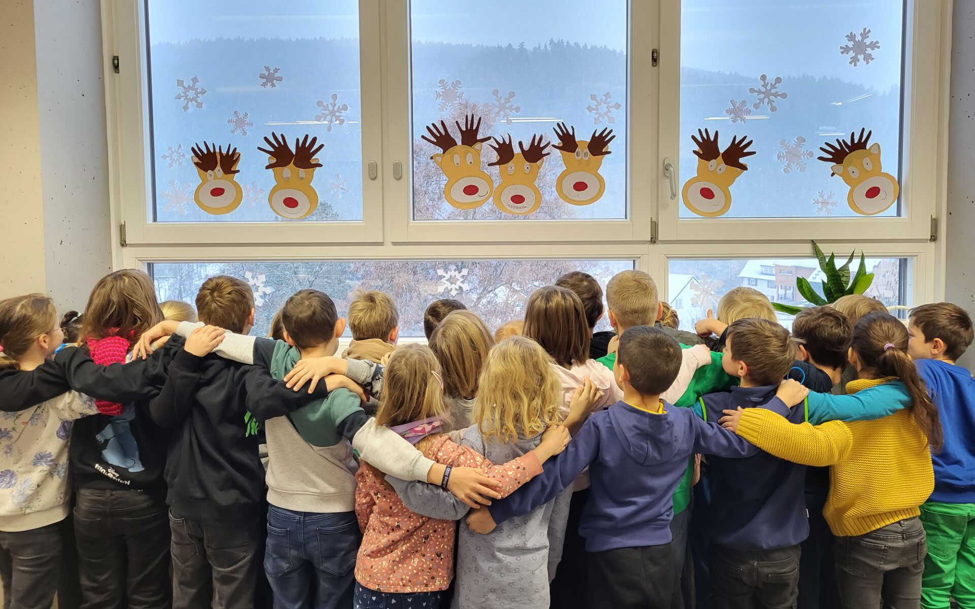 Die Kinder der ersten Klasse stehen am Fenster und blicken hinaus. Einige Kinder umarmen sich. Das Fenster ist winterlich dekoriert.