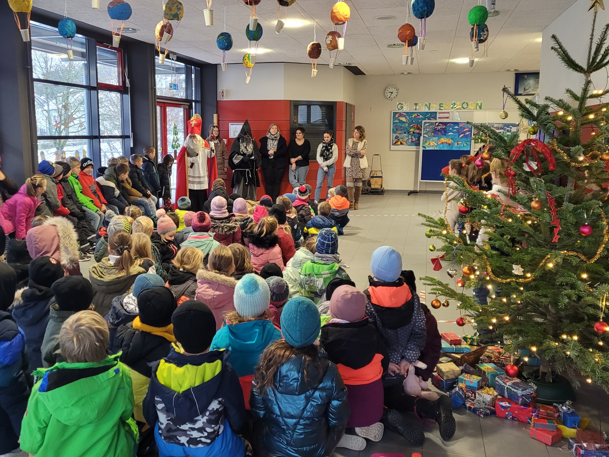 Viele Kinder sitzen in der Pausenhalle neben einem Weihnachtsbaum. Im Hintergrund sind Nikolaus, Knecht Ruprecht und einige Lehrerinnen zu sehen.