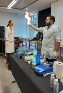 Ein Mann in Laborkleidung hält seine brennende Hand in die Höhe. Im Hintergrund ist eine lächelnde Frau in Laborkleidung zu sehen.