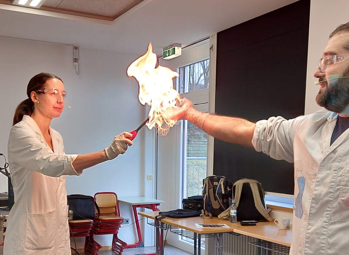 Eine Frau in Laborkleidung zündet hält ein Feuerzeug an die Hand eines Mannes. Die Hand steht komplett in Flammen