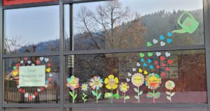 "Schulgarten der Freundlichkeit": Ein aus Papier gebasteltes Fensterbild, auf dem Blumen aus einer Gießkanne mit Herzen gegossen werden