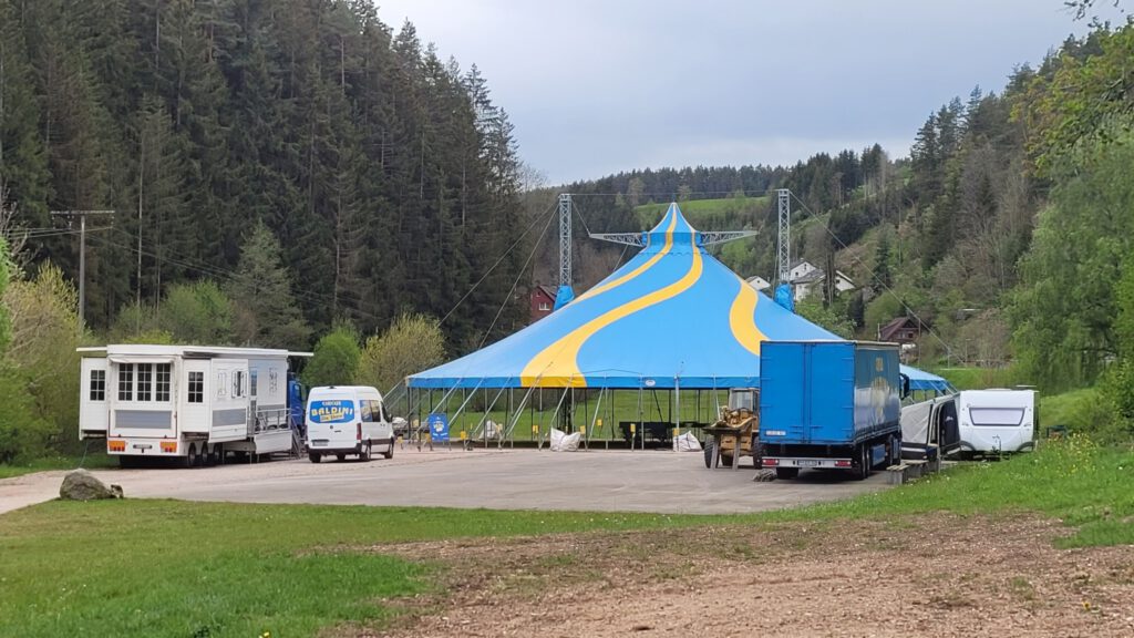 Auf einem großen Platz ist in der Mitte das Dach eines Zirkuszeltes zu sehen, die Seitenplanen fehlen noch. Rechts und links des Zeltes stehen Wohn- und Lastwagen.